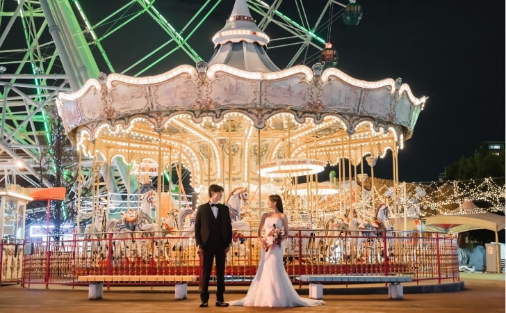 Nagoya Port Sea Train Land｜洋装姿のカップルが夜の遊園地でメリーゴーランドを背景に撮影をしているイメージ
