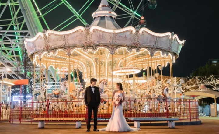 Nagoya Port Sea Train Land｜洋装姿のカップルが夜の遊園地でメリーゴーランドを背景に撮影をしているイメージ